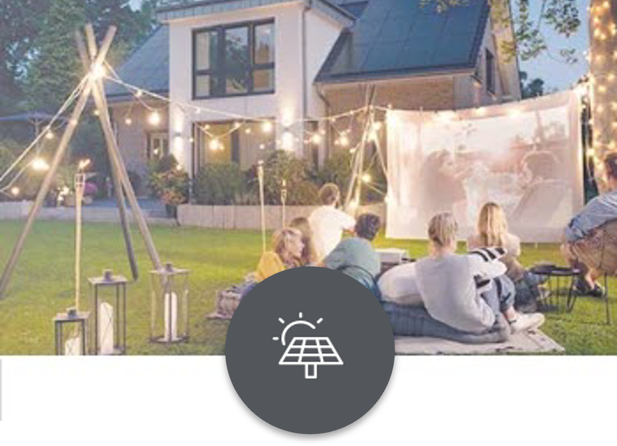 Solaranlage Eine Familie sitzt gemütlich auf dem Rasen auf einer Decke und schaut einen Film auf einer Projektionsleinwand. Sie sind umgeben von dekorativen warmweißen Lichterketten. Im Hintergrund ein modernes Haus mit einer Solaranlage auf dem Dach.